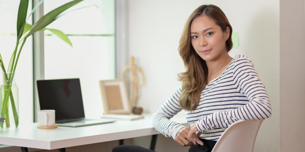 Femme souriante au bureau : espace de travail moderne et lumineux. Jeune femme souriante, chemise rayée, assise au bureau. Espace de travail lumineux avec ordinateur portable, mug, plante et figurine en bois.