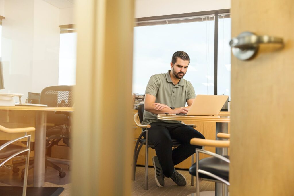 Travail au bureau : homme concentré sur son ordinateur portable. Homme concentré tapant sur son ordinateur portable dans un bureau clair, vu à travers une porte ouverte. Lumière naturelle.