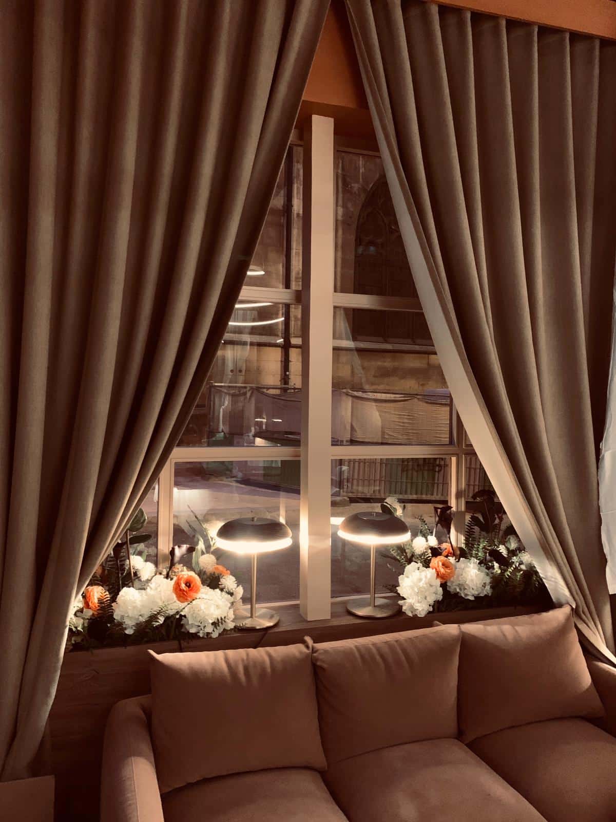 Intérieur cosy. Canapé velours, rideaux marrons. Lumières douces sur des fleurs. Vue nocturne sur une fenêtre gothique en pierre.