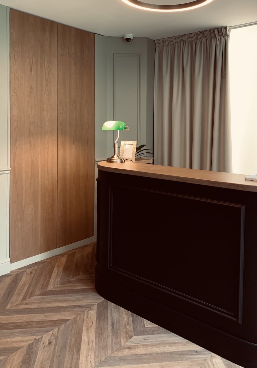 Comptoir de réception noir incurvé avec lampe de banquier verte, armoires en chêne clair et sol en parquet à chevrons élégant.