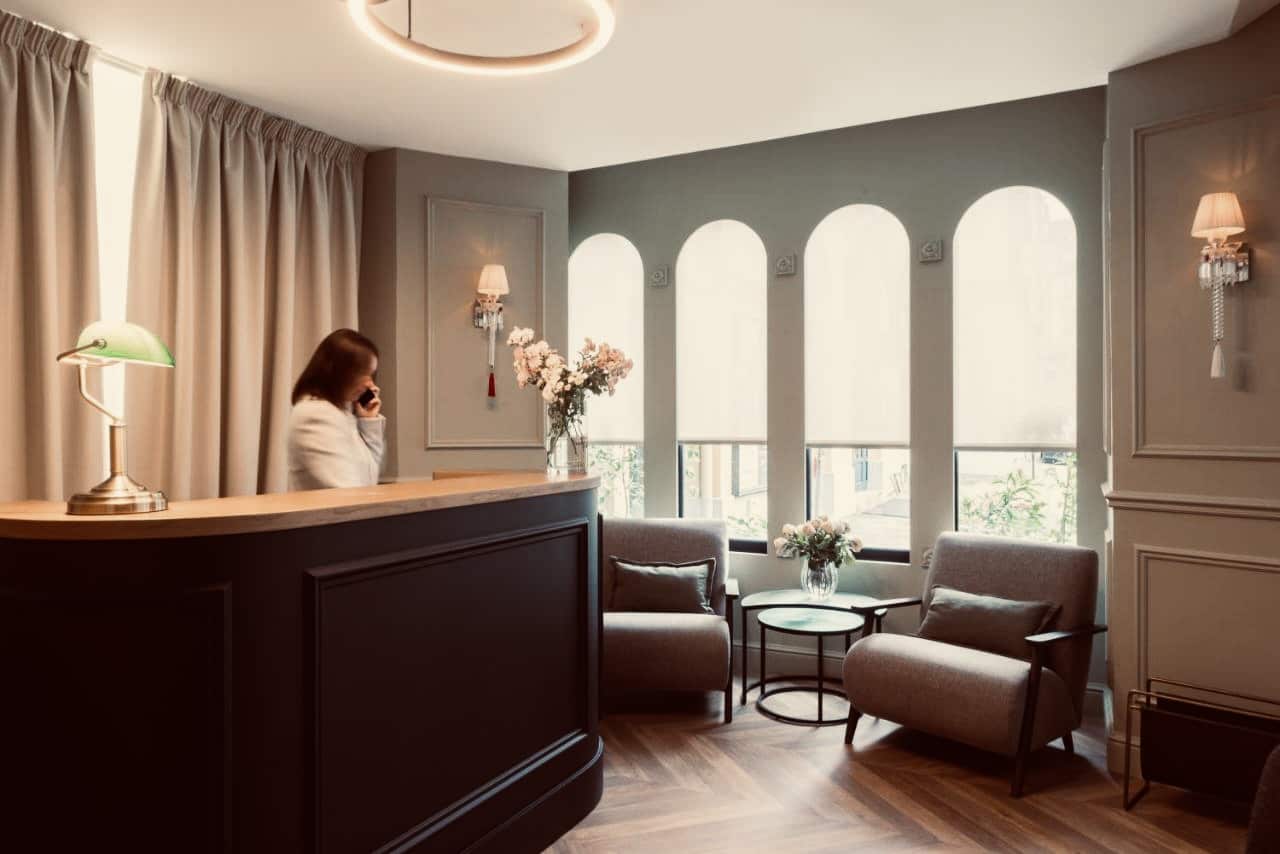 Lobby d'hôtel raffiné. Réceptionniste au téléphone derrière un comptoir noir. Décor élégant avec moulures, lampe de banquier et fauteuils près des fenêtres arquées.