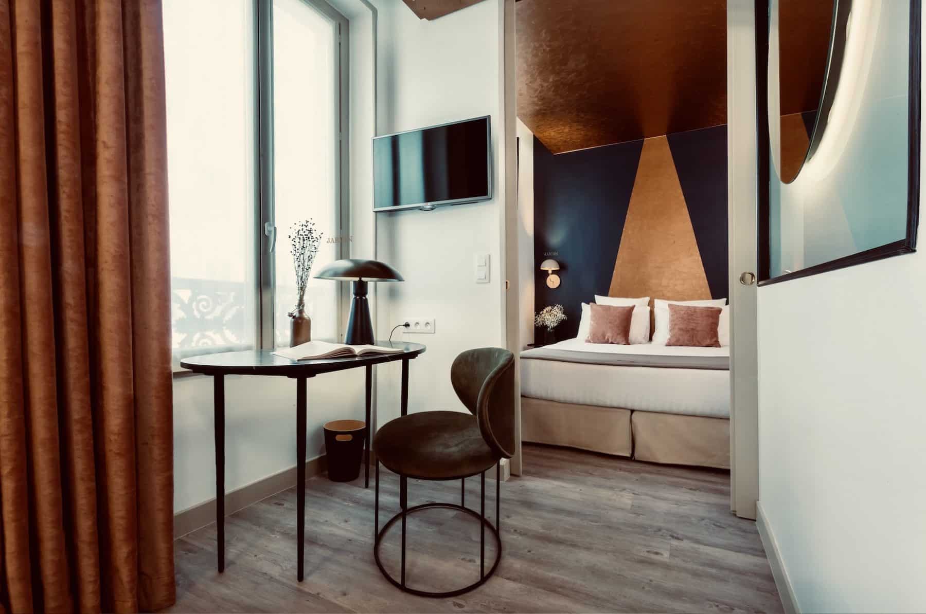 Chambre d'hôtel design luxe. Bureau avec chaise velours vert près d'une fenêtre inscrite JARDIN. Au fond, lit avec tête de lit géométrique bleu et cuivre.