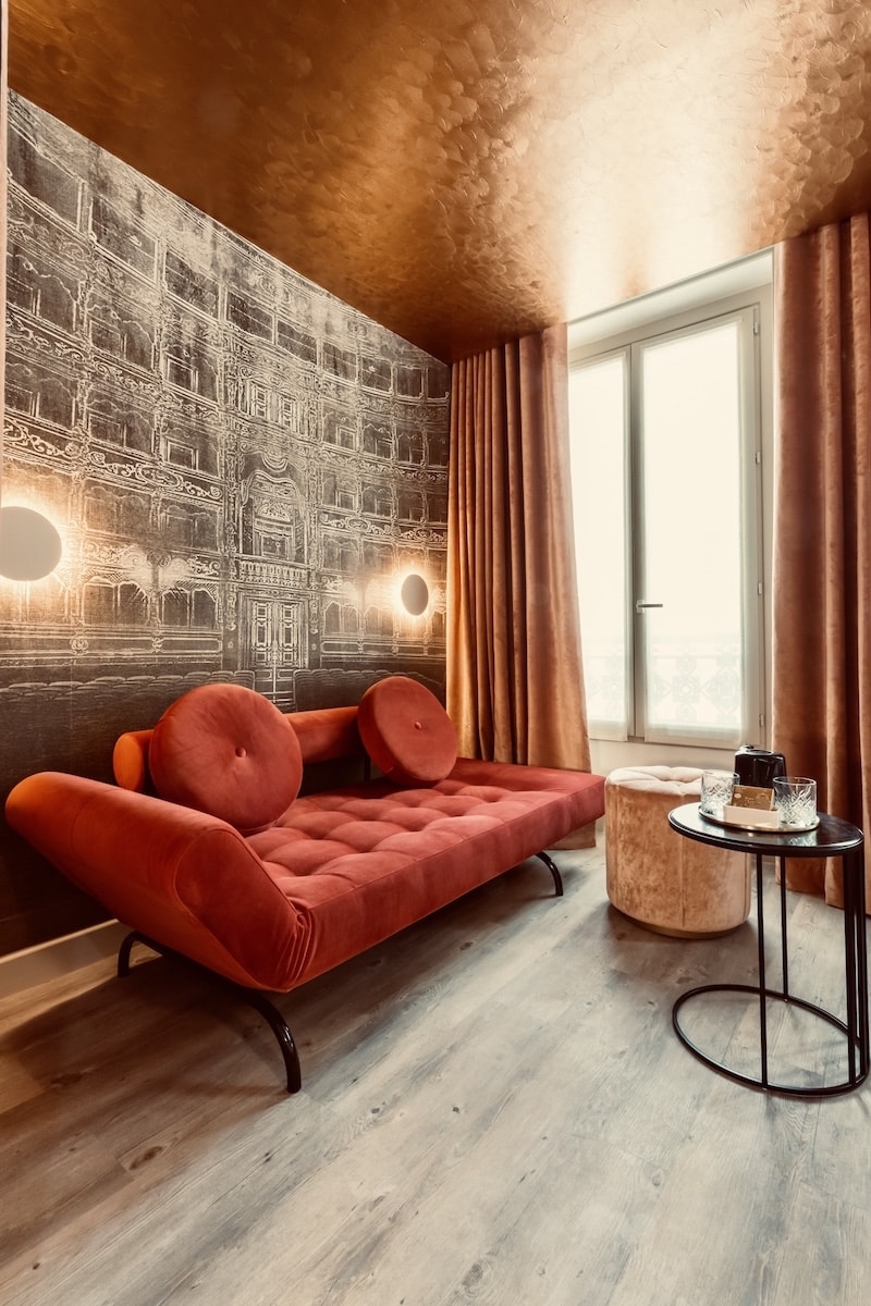 Somptueux daybed en velours rouge devant un papier peint architectural sombre. Plafond en cuivre martelé et rideaux terracotta.