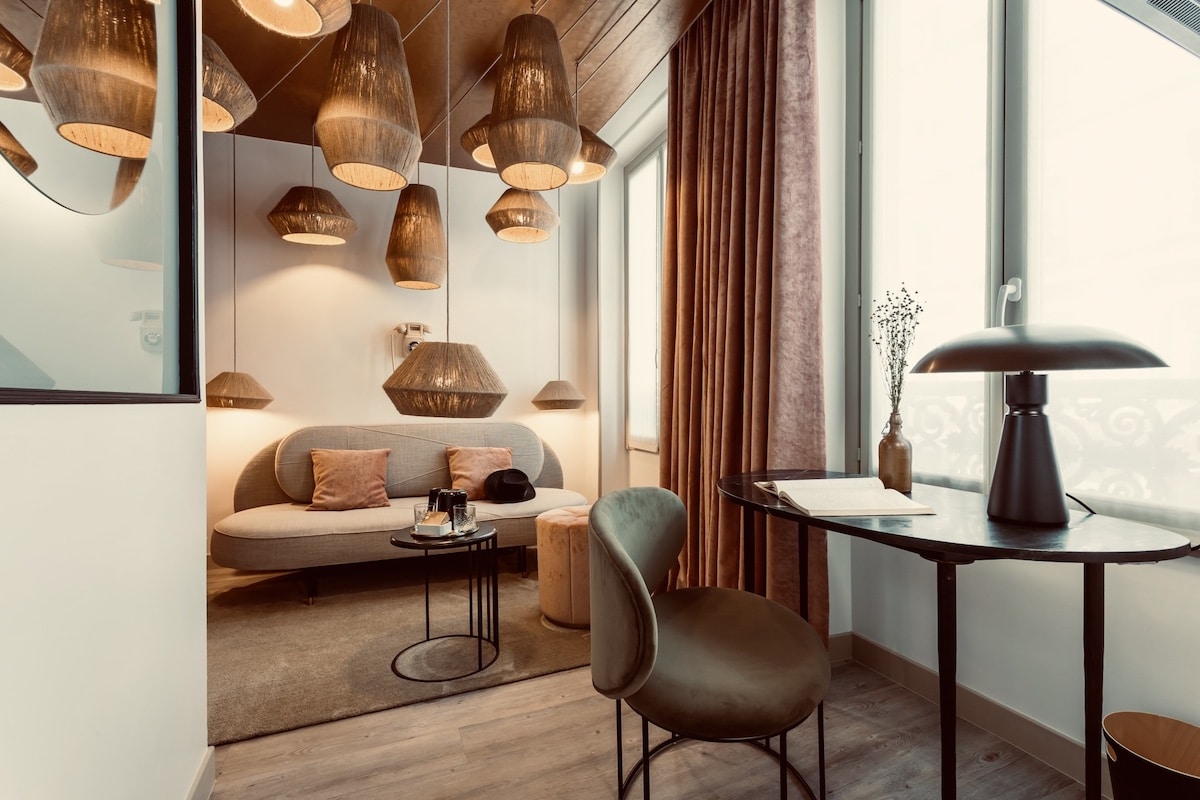 Salon chic avec multiples suspensions tissées, canapé arrondi et coussins roses. Chaise en velours vert et lampe de bureau design.