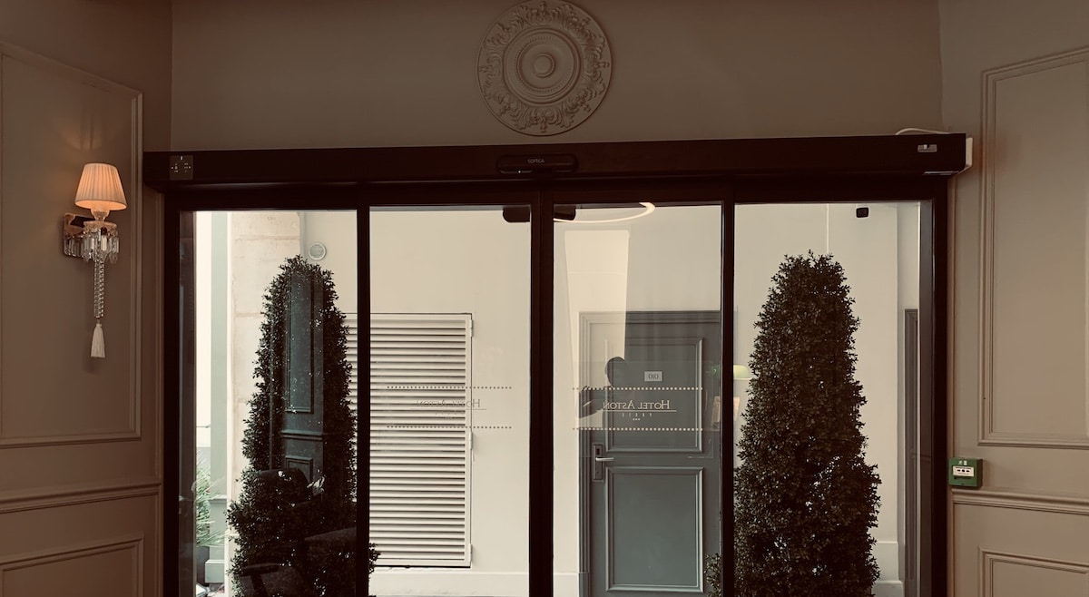 Entrée chic de l'Hotel Aston. Porte vitrée automatique Softica. Murs taupe moulurés, sconce en cristal et deux topiaires vertes.