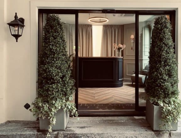 Entrée vitrée de l'Hôtel Aston Paris. Réception noire, parquet chevron, lumière moderne, et deux élégants topiaires flanquant l'accès.