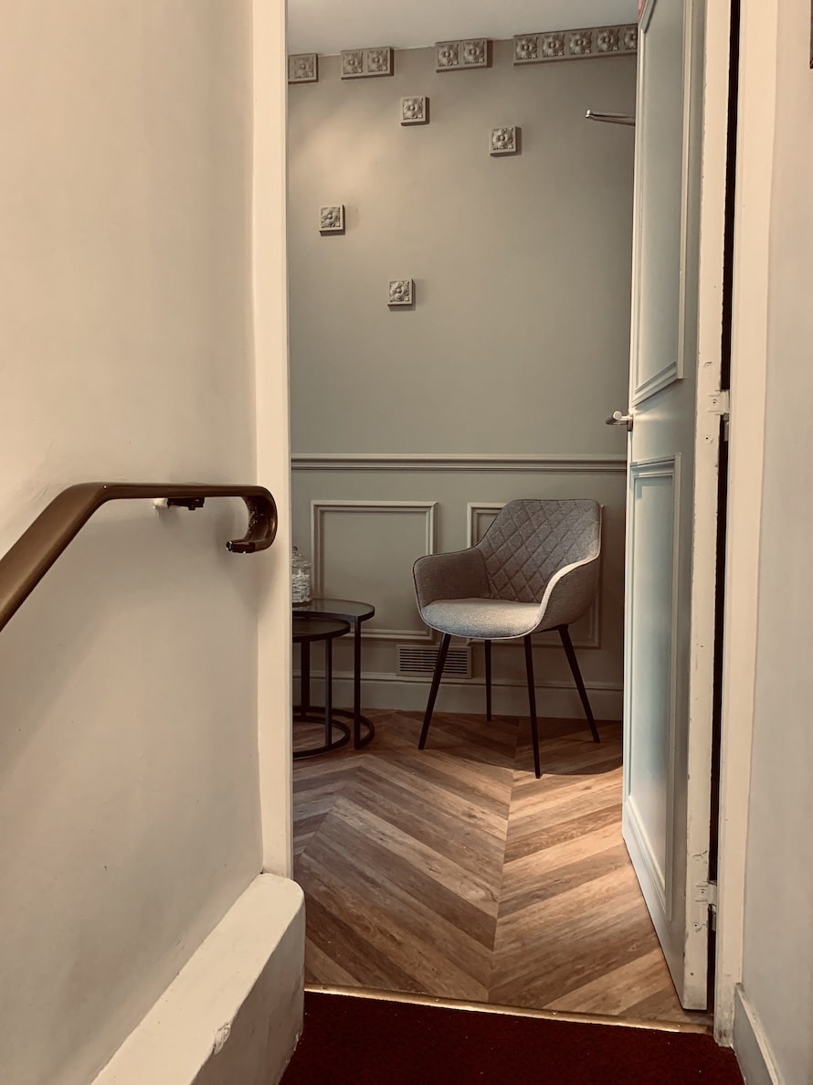 Fauteuil gris dans une salle d'attente élégante. Boiseries gris-vert, décor mural de moulures et sol en parquet chevron.