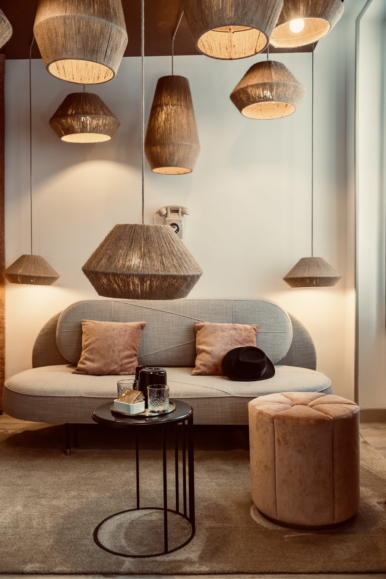 Salon cosy: canapé design, pouf velours rose, table d'appoint noire et multiples suspensions en jute éclairant un téléphone vintage au mur.