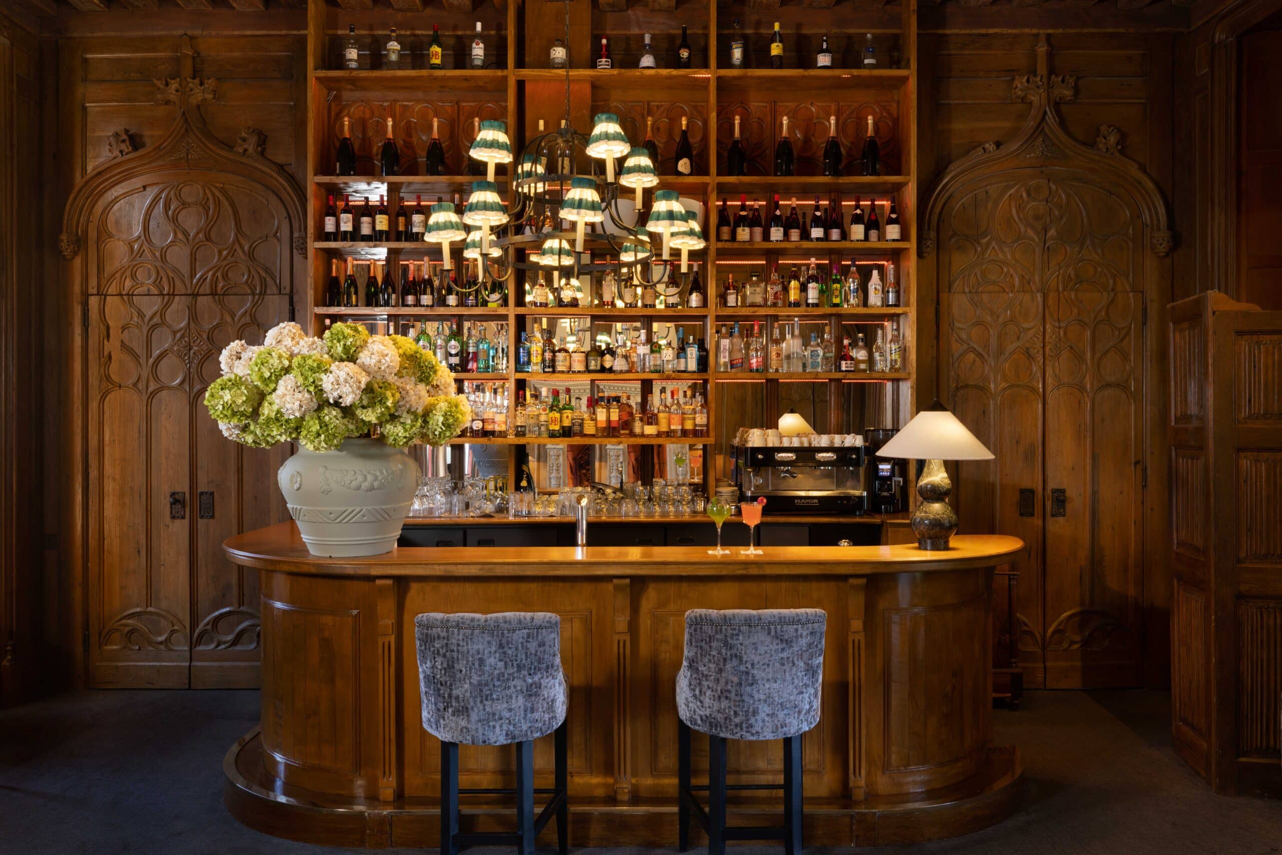 Élégant bar en chêne sculpté (boiserie). Étagères illuminées de champagne et spiritueux, lustre vert. Tabourets et cocktails sur le comptoir.