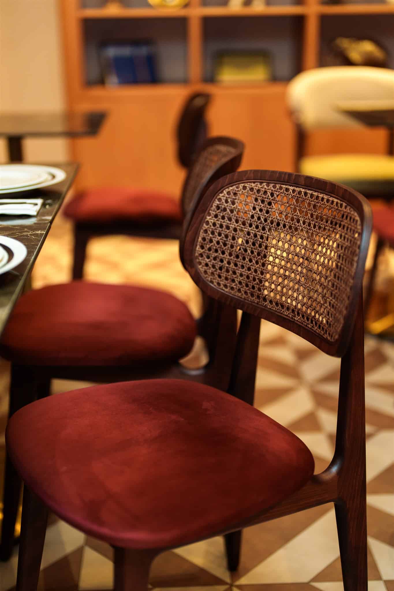 Chaise de restaurant en bois foncé avec dossier en cannage et siège en velours bordeaux. Table en marbre et étagères en arrière-plan flou.
