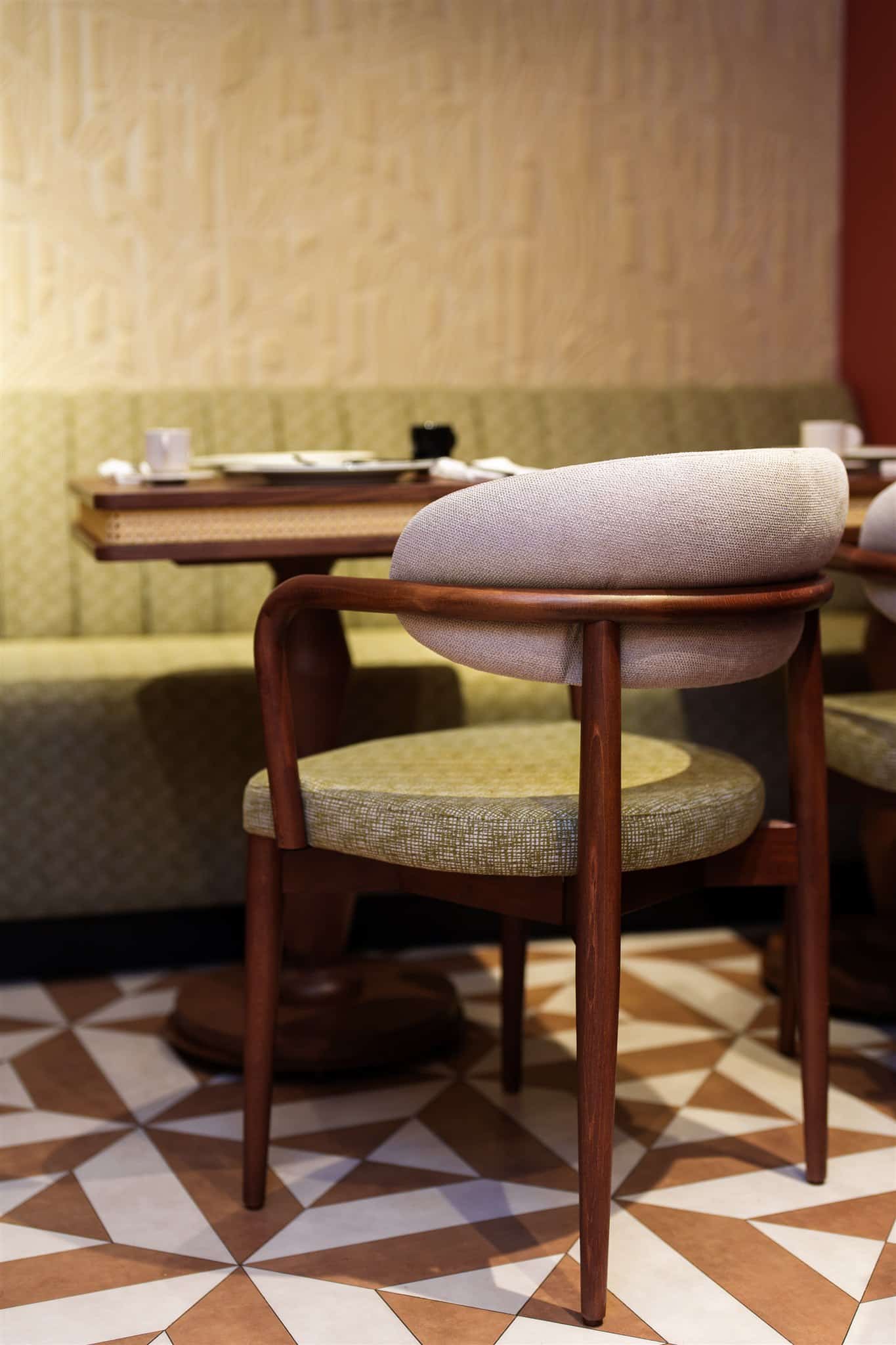 Chaise de restaurant bois et tissu, dossier beige, assise verte. Banquette olive, table dressée et mur texturé clair. Sol à carrelage géométrique marron.