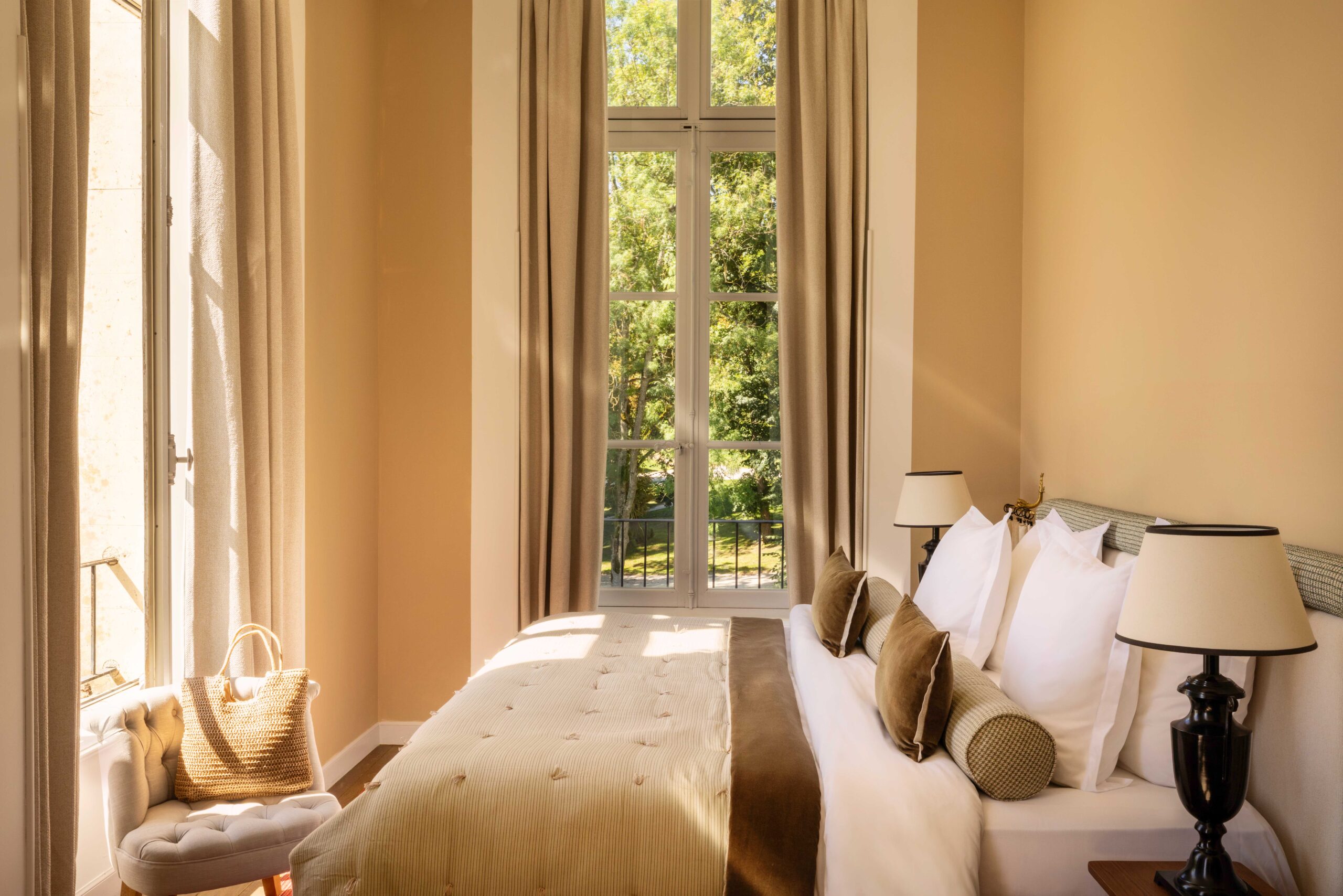 Chambre d'hôtel lumineuse et élégante. Lit double beige, murs taupe, et fauteuil tufté. Belle vue sur un parc verdoyant à travers la fenêtre.