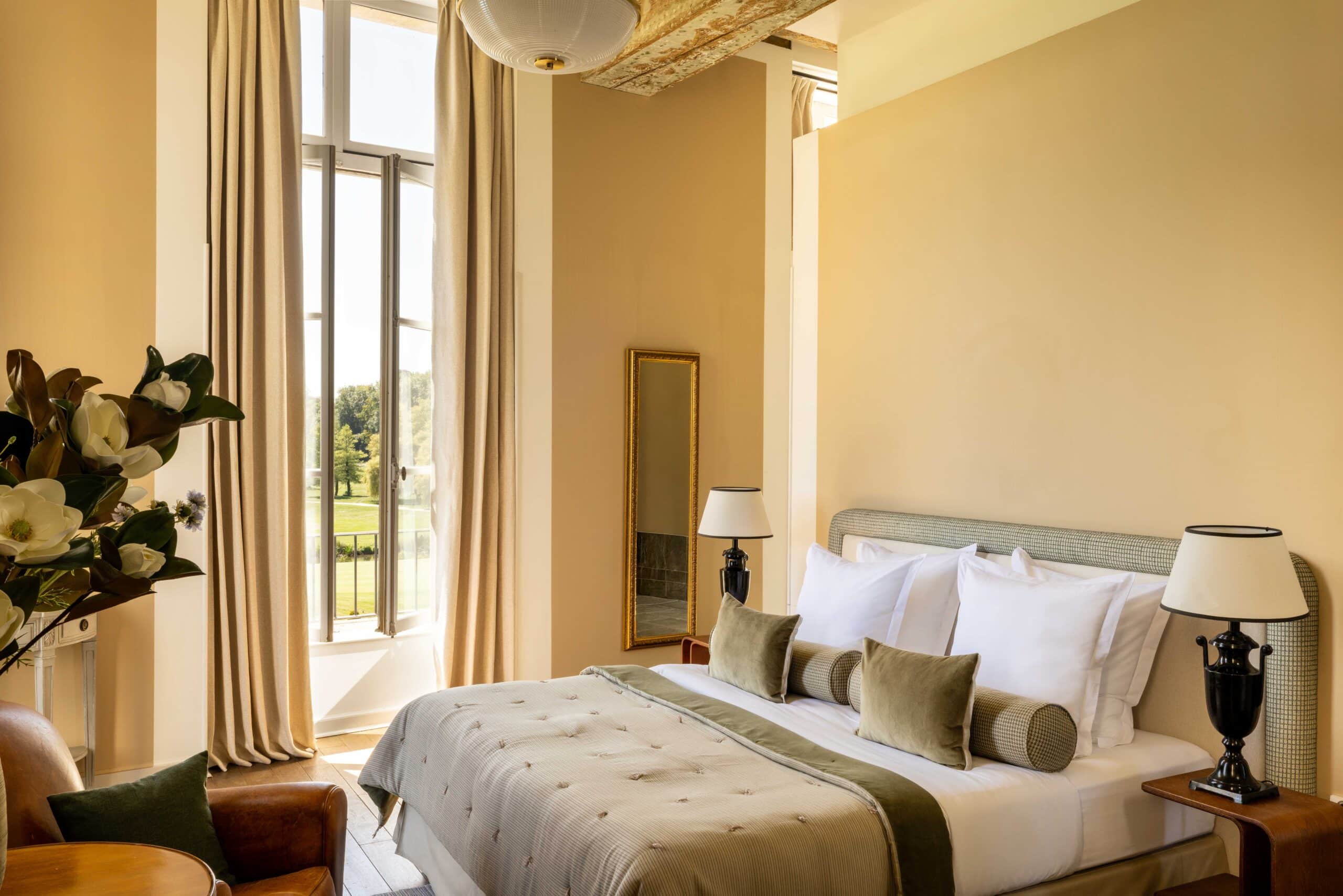 Luxueuse chambre d'hôtel aux murs beige chaud. Grand lit décoré de vert sauge. Vue imprenable sur un parc par la haute fenêtre.
