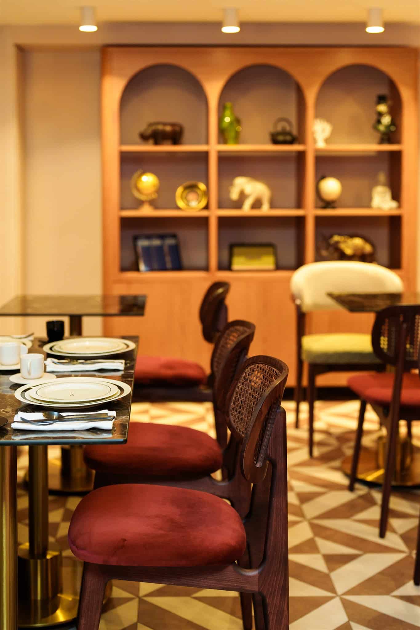 Espace repas élégant : chaises en bois canné, coussins velours rouge. Table dressée devant des étagères murales arquées et décorées.