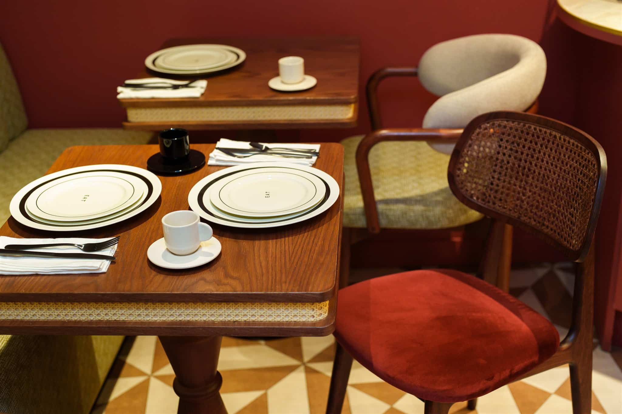 Table de restaurant en bois. Assiettes blanches marquées "EAT", couverts. Chaise rétro en rotin et velours rouge. Décor intérieur cosy et riche.