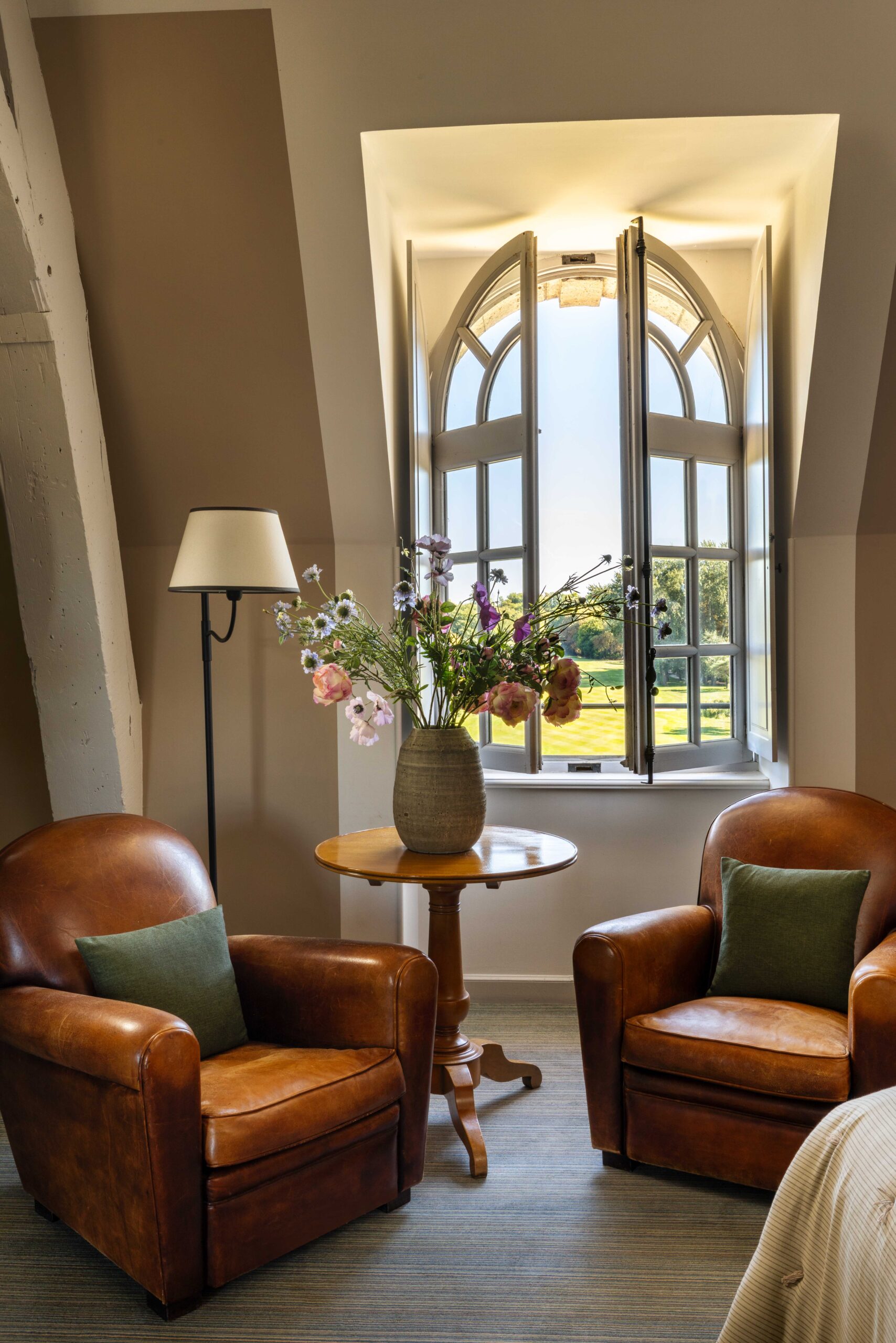 Coin salon mansardé avec deux fauteuils club en cuir marron. Des fleurs fraîches sur la table devant une grande fenêtre arquée donnant sur le parc.