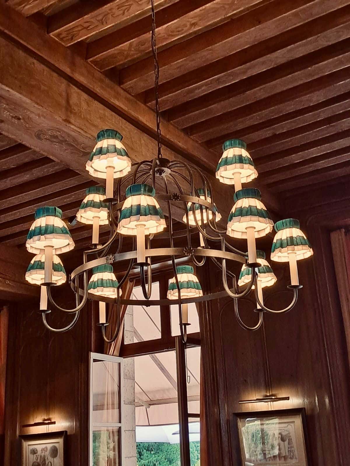 Ornate lustre suspendu en fer forgé. Ses nombreux abat-jour rayés (vert et blanc) éclairent les boiseries sombres et le plafond à poutres.
