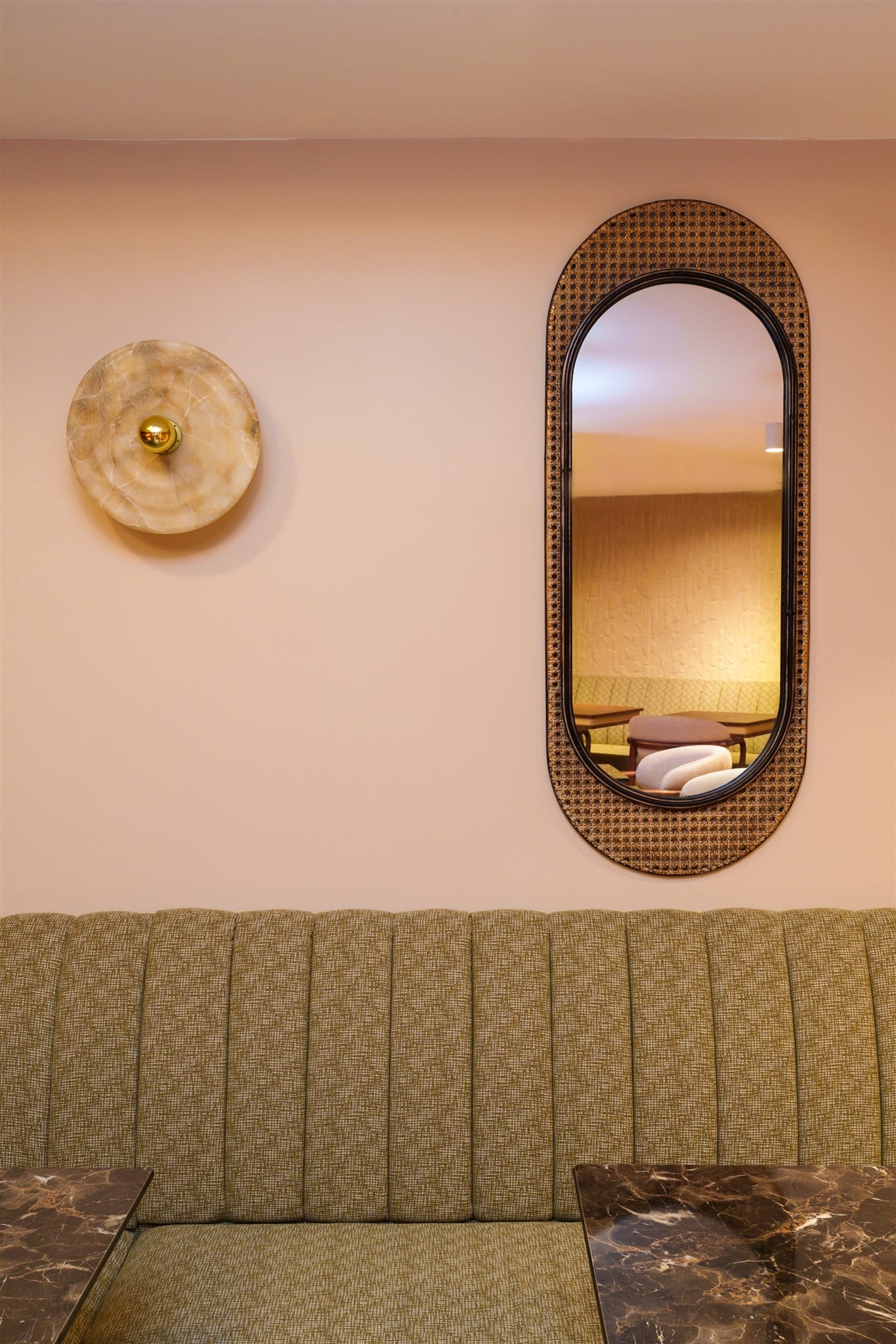Banquette vert olive flutée sous un mur rose. Applique murale en onyx et miroir ovale en cannage encadré de noir.