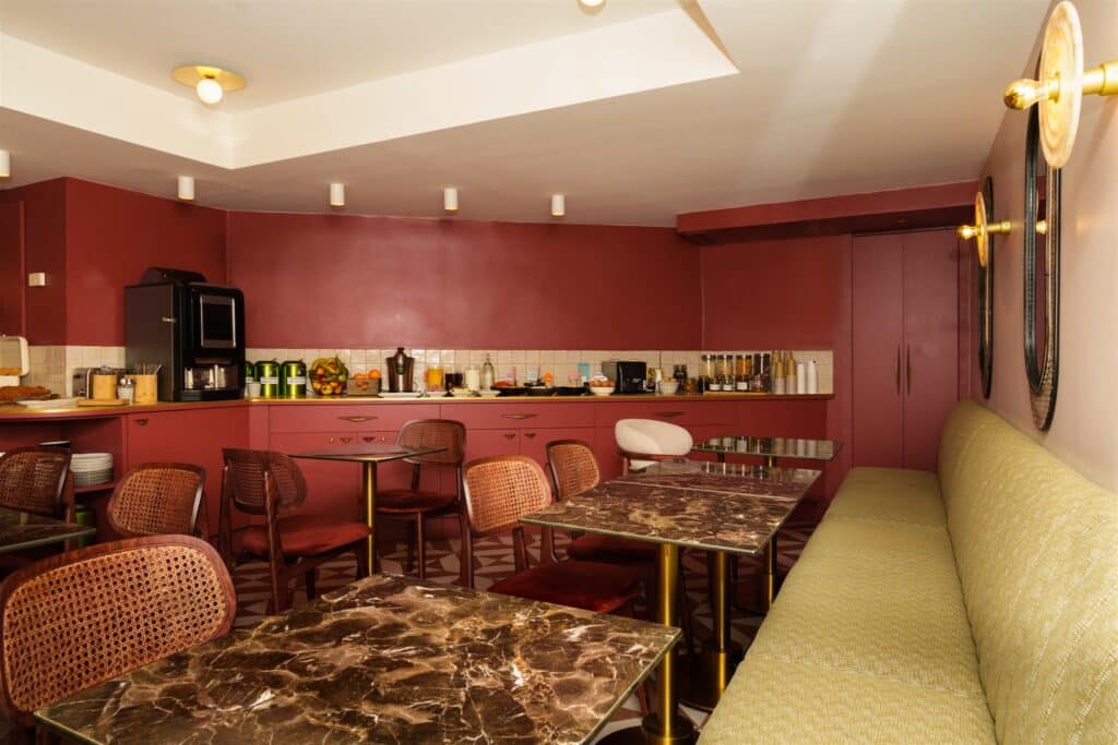 Salle de petit-déjeuner rétro chic : murs bordeaux, buffet garni, chaises rotin et banquette vert olive. Tables marbre et laiton.