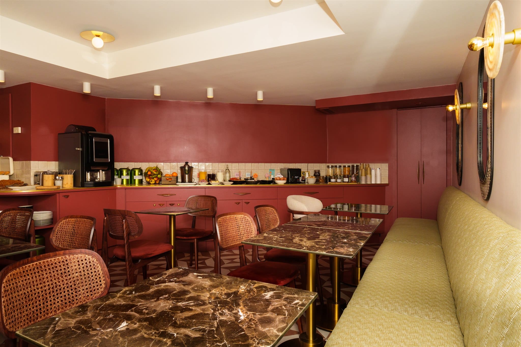 Salle de petit-déjeuner rétro chic : murs bordeaux, buffet garni, chaises rotin et banquette vert olive. Tables marbre et laiton.