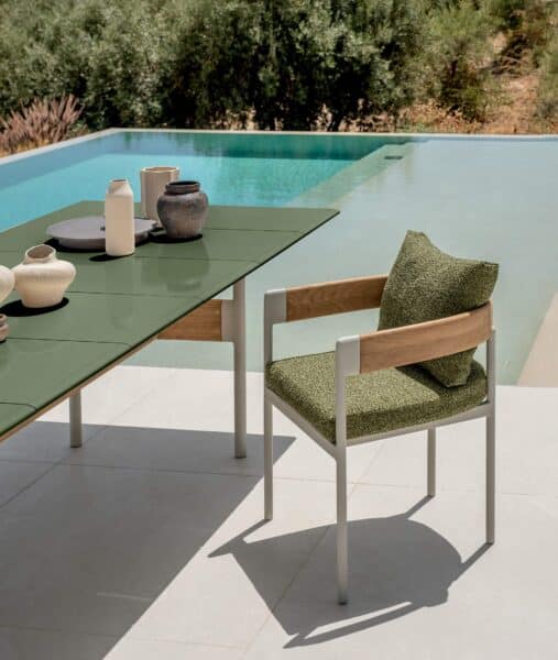 Table à manger extérieure vert olive et chaise design avec accoudoirs en bois, cadre gris et coussin vert, près d'une piscine.
