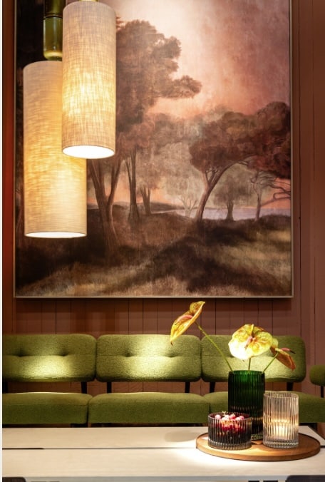 Sofa vert olive et grand tableau de forêt sombre sur mur terracotta. Suspensions en lin chaud et vases nervurés sur plateau de table.