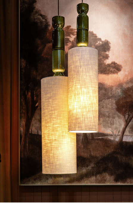 Deux suspensions lumineuses: céramique vert foncé et abat-jours cylindriques en lin. Ambiance chaleureuse devant une peinture de forêt.